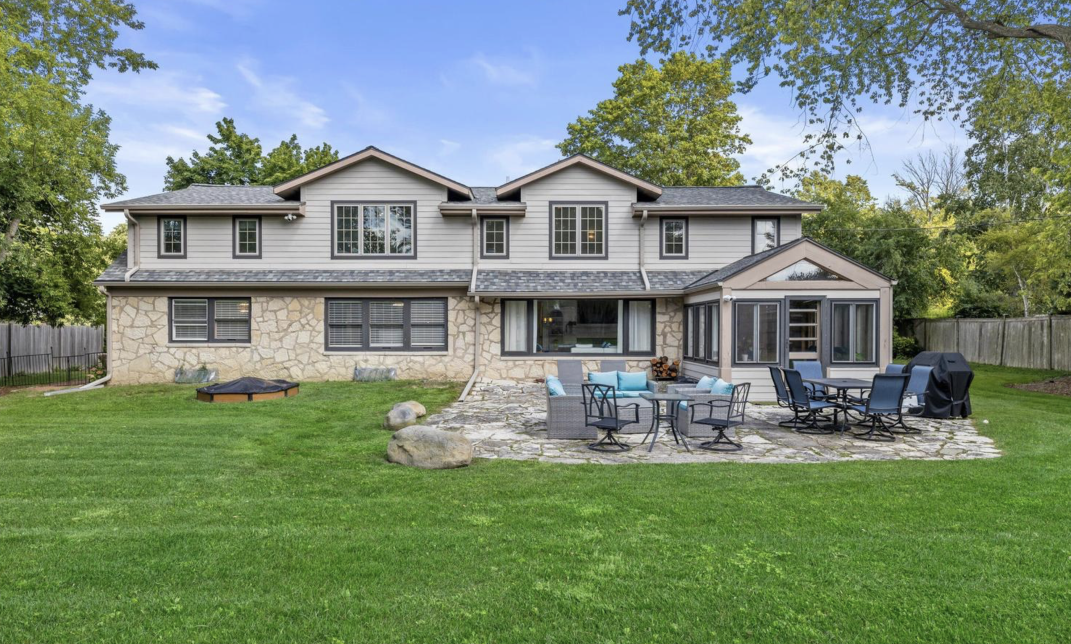 Backyard with green grass, a stone patio with outdoor furniture, a grill, trees, and a two-story house with large windows.