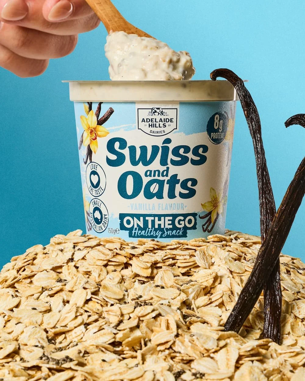 A cup of Swiss and Oats vanilla flavor yogurt being scooped out with a wooden spoon, surrounded by oats and vanilla beans, against a light blue background.