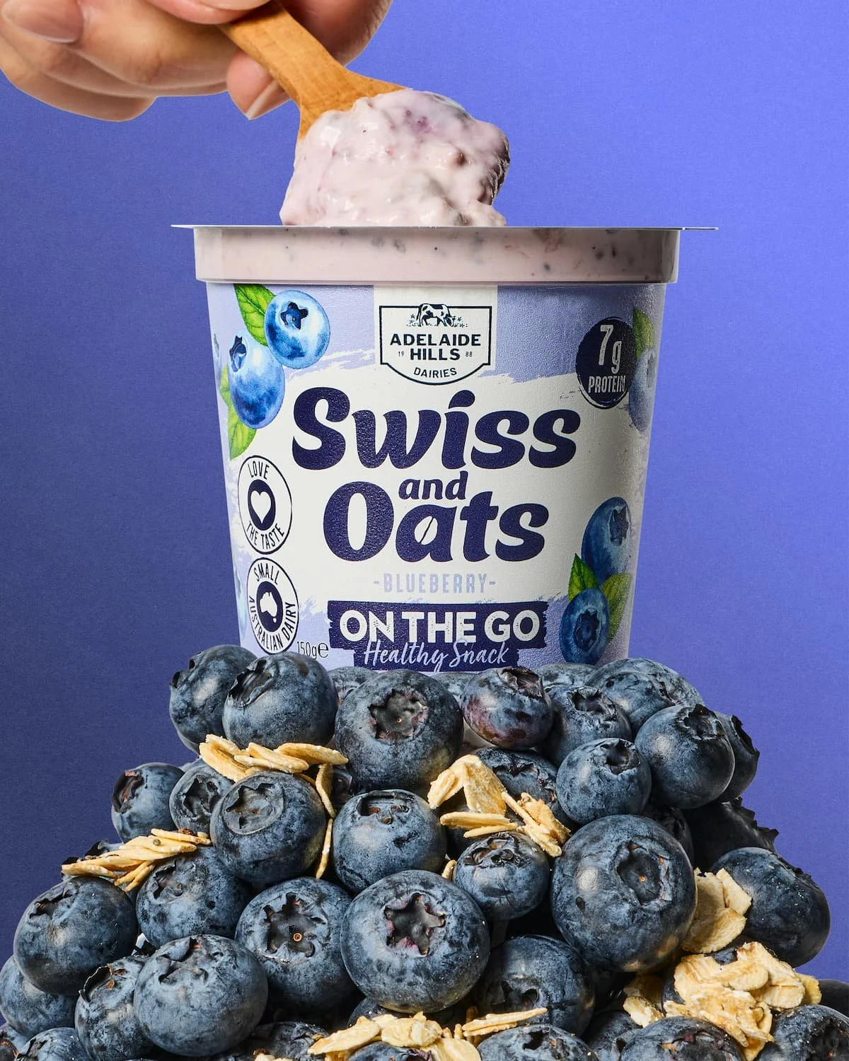 A container of Swiss and Oats blueberry flavored on-the-go snack surrounded by fresh blueberries and oat flakes.