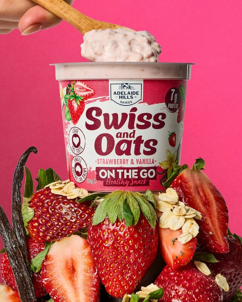 A tub of Adelaide Hills Swiss and Oats strawberry and vanilla flavored yogurt on a pink background, surrounded by fresh strawberries, oats, and a vanilla bean.