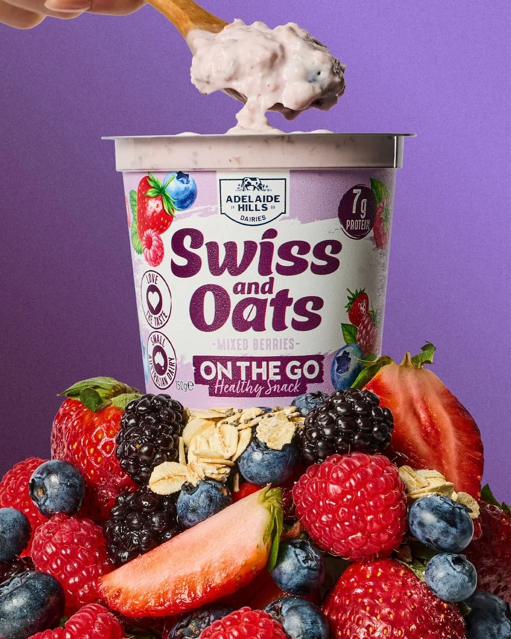 A container of Adelaide Hills Swiss and Oats mixed berries yogurt on a purple background, surrounded by fresh strawberries, blackberries, blueberries, and some granola.