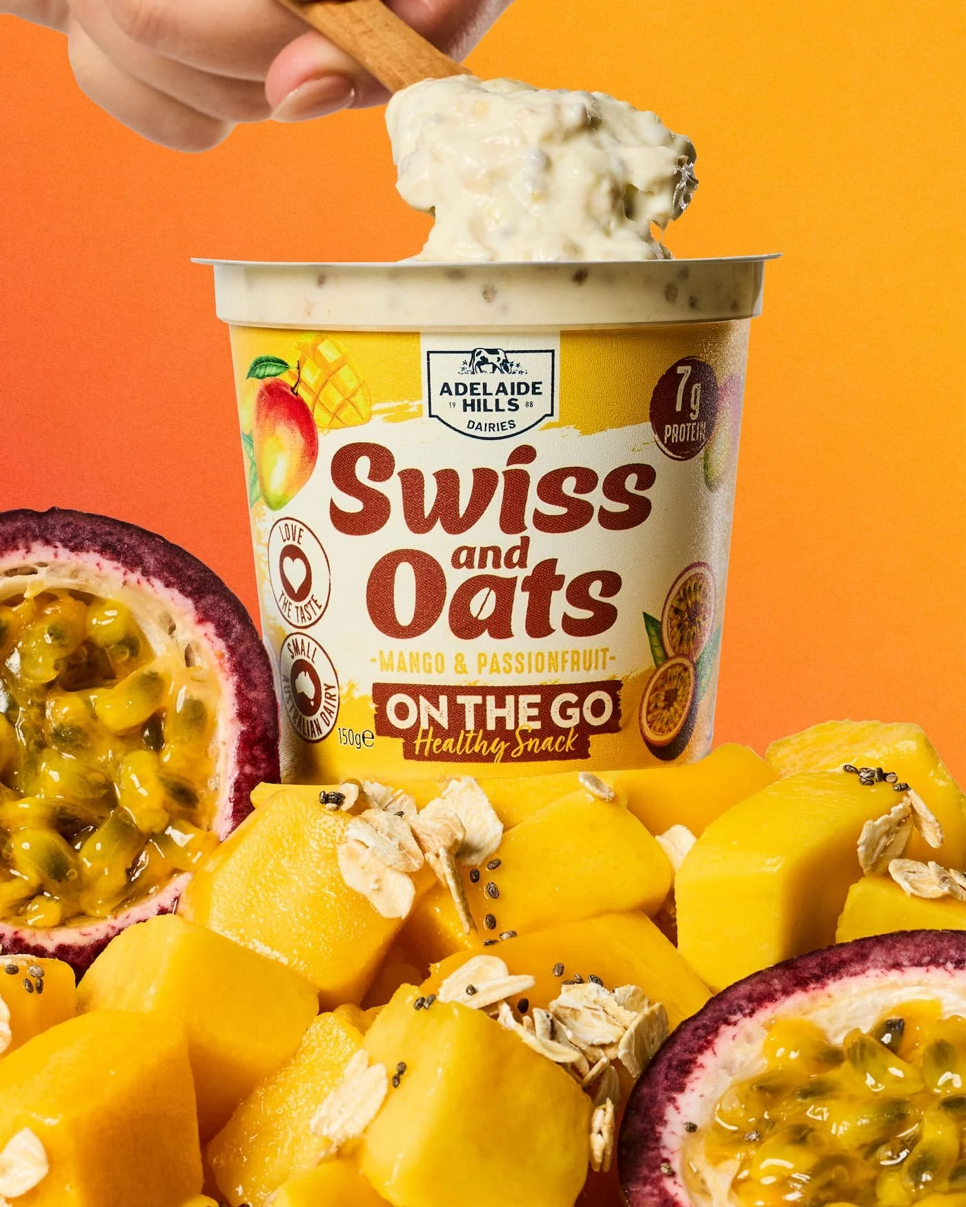 A container of Swiss and Oats Mango & Passionfruit snack with passionfruit halves and diced mango pieces around it, against a orange background.