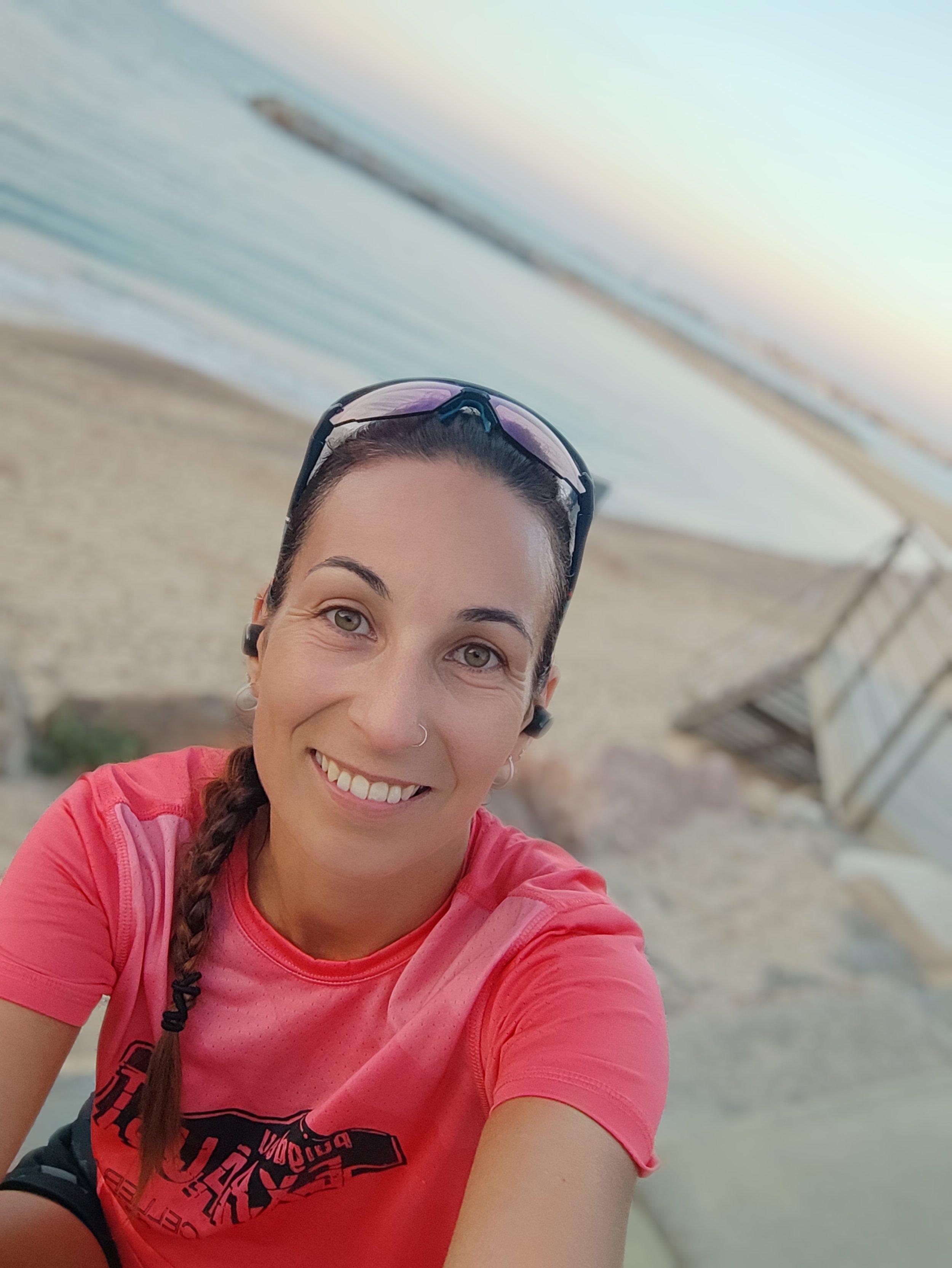 Mujer sonriendo en la playa, usando gafas de sol, con cabello en trenza y camiseta color rosa, fondo de arena y agua.