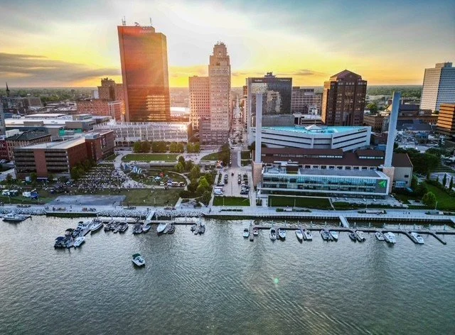 Aerial view of a city skyline at sunset with high-rise buildings, a waterfront, and boats docked along the shore.