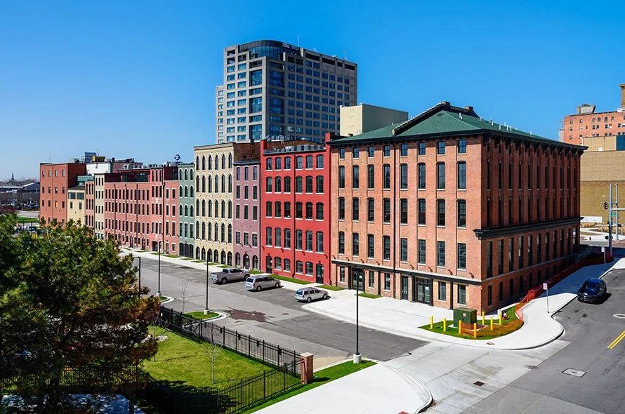 Colorful row of historic brick townhouses with parked cars in front, modern high-rise building in the background, blue sky, and a small fenced grassy area with trees in the foreground.