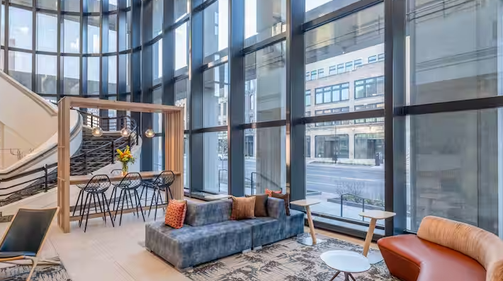 Modern hotel lobby with large glass windows, contemporary sofas, chairs, and a staircase with black railing and wooden accents.