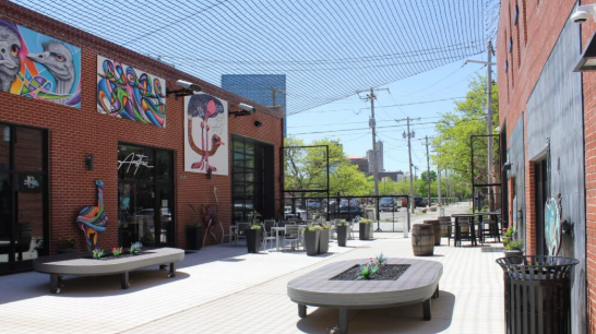 Outdoor urban patio with colorful murals on brick walls, seating, planters, and trash bins, with city buildings and trees in the background.
