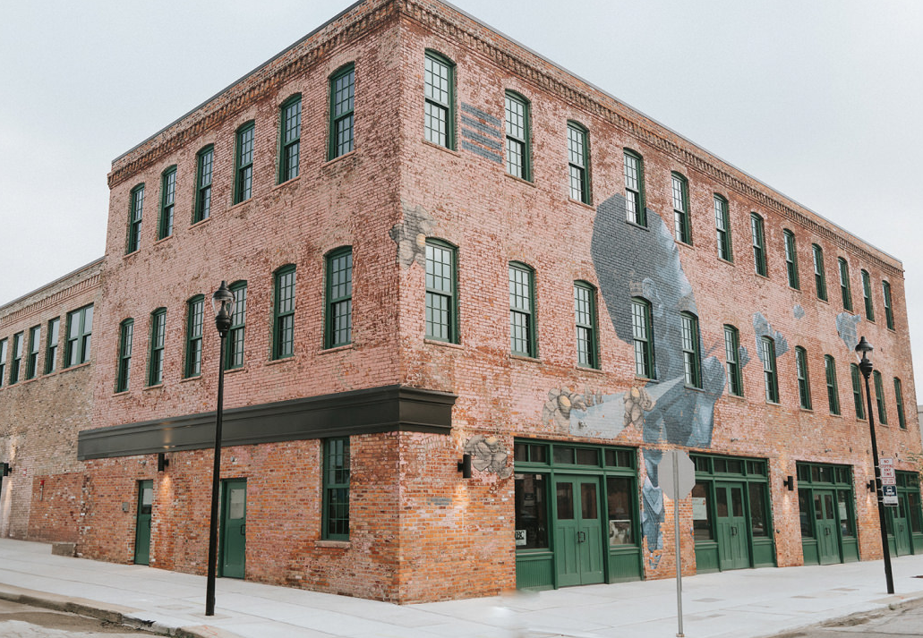 A multi-story brick building with green window frames and doors, decorated with a large mural of a woman wearing a blue hooded garment on the side wall, on a city sidewalk.