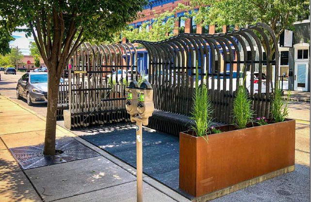 Public parking meter, decorative wooden shelter with plants, sidewalk, parked cars, trees in an urban street scene.