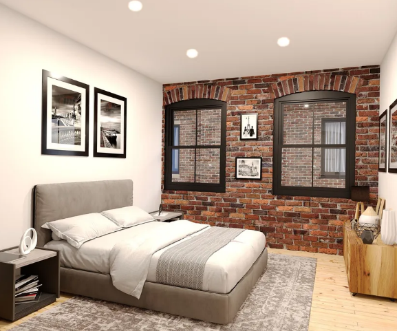 Modern bedroom with a gray upholstered bed, white bedding, framed black and white photographs on the wall, two black-framed windows revealing brick walls outside, a wooden dresser with decorative objects, and a patterned rug on light wood flooring.