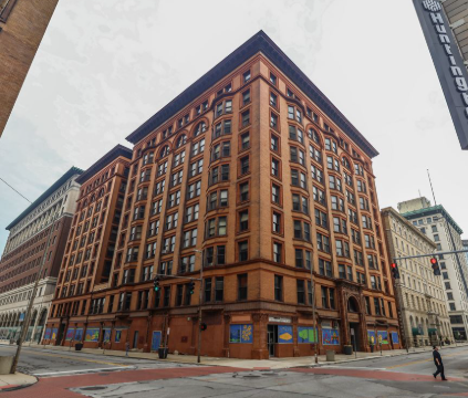 A tall brick building on a city street corner, with multiple windows and a flat roof, surrounded by other buildings and a few people walking by.
