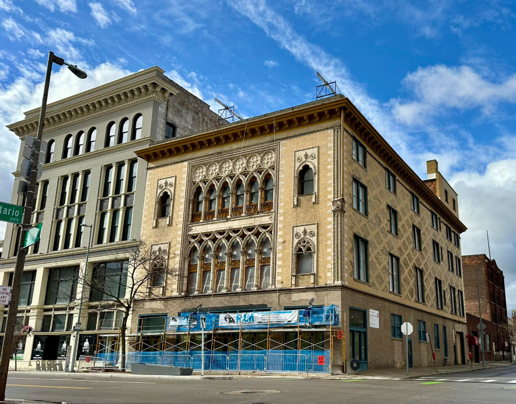 City street scene with historic building featuring ornate windows and detailed architectural design, partly under construction with scaffolding and blue barriers, under a blue sky with some clouds.