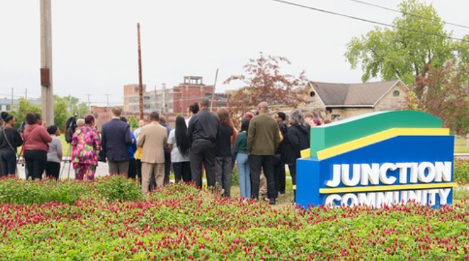 A group of people gathered outdoors near a colorful community sign that says 'Junction Community,' with greenery and trees in the background.