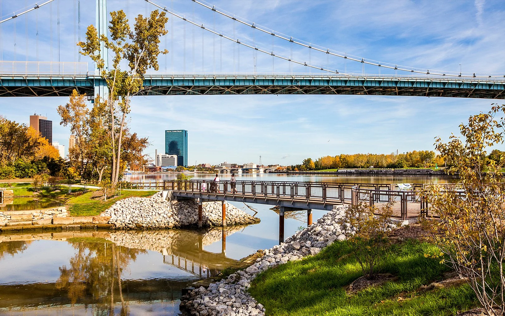City park overlooking a river with walking paths, trees in fall, and bridges, with city buildings in the background.