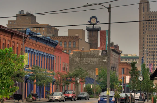 Street scene with colorful buildings, parked cars, trees, utility poles, and a clock on a building in an urban setting.
