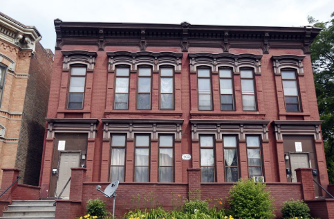 Red brick building with three stories, large windows, and greenery in front.
