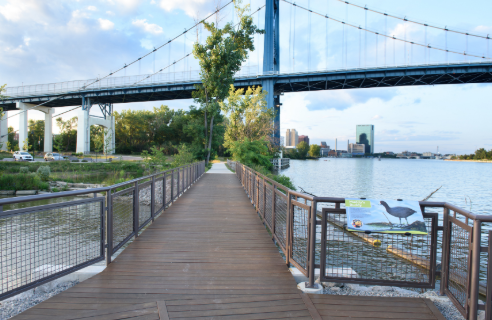 Wooden walkway along a river with a bridge overhead, trees, and city skyline in the background.