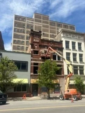 Building under construction with a crane in front, trees, and parked cars along the street.