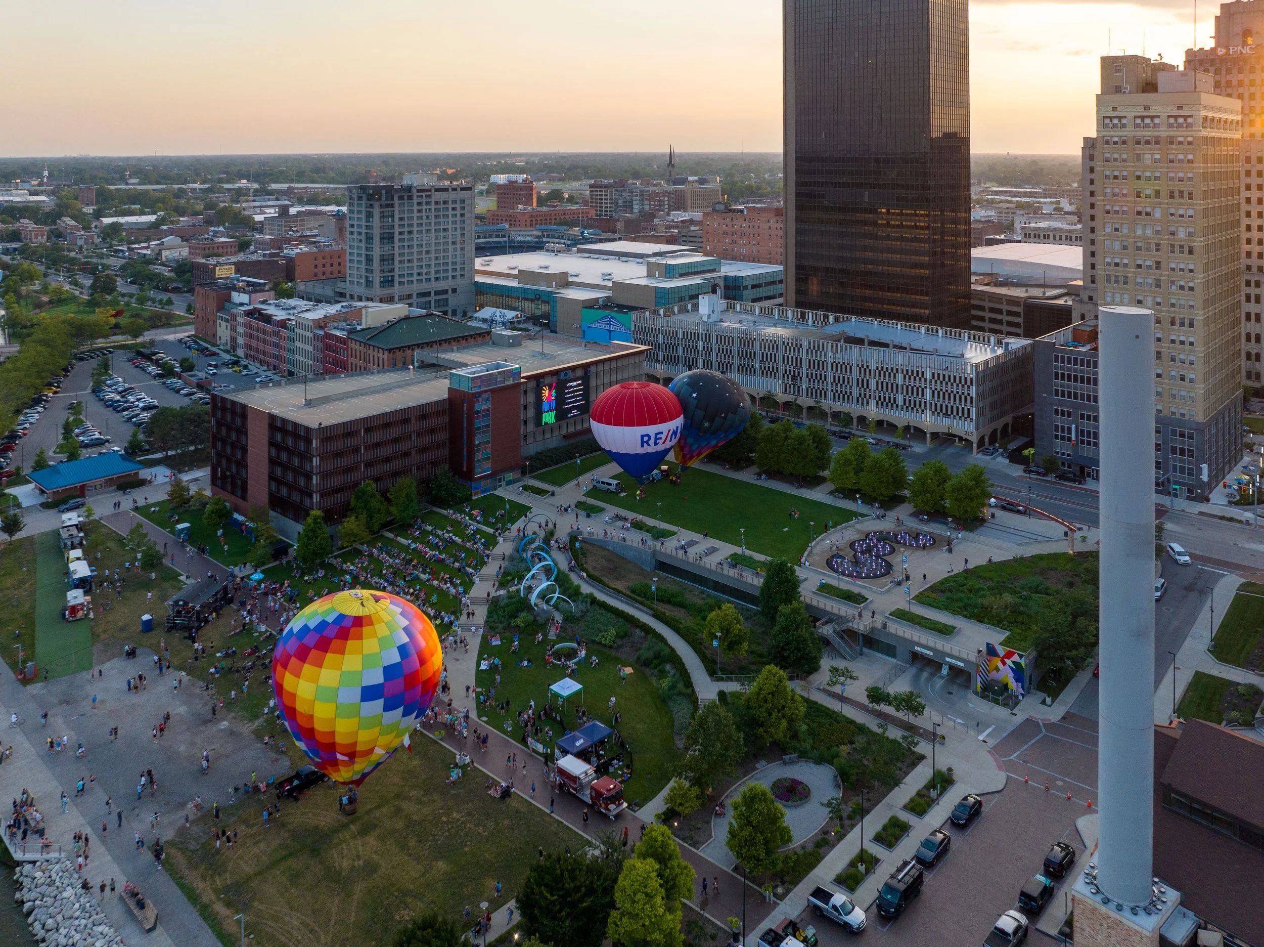 Aerial view of a city park during a hot air balloon event at sunset, with colorful balloons, numerous visitors, and surrounding city buildings.