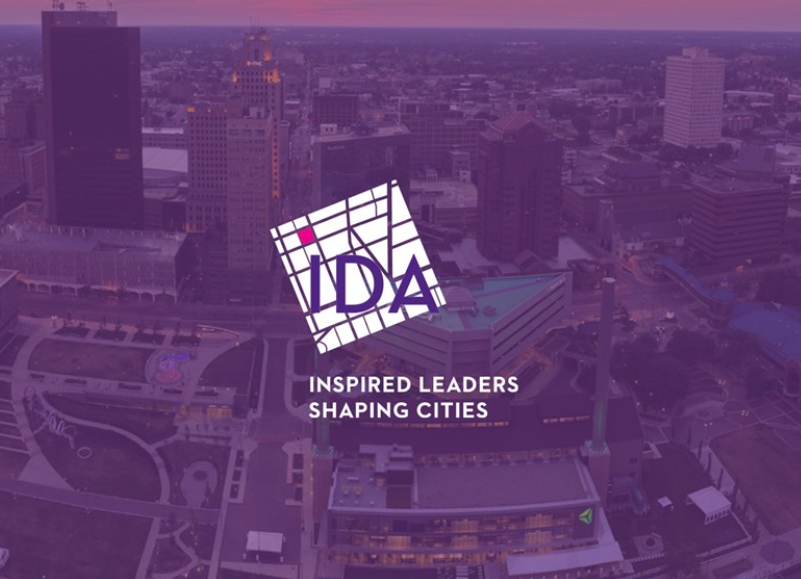 Aerial view of a cityscape with tall buildings at sunset, featuring a logo for IDA and the slogan 'Inspired Leaders Shaping Cities' overlayed on the image.