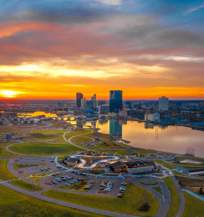 A city skyline at sunset with tall buildings reflecting on a river, and a park with walking paths and parking lots in the foreground.