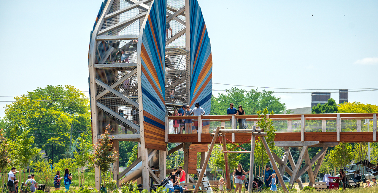 A large outdoor playground structure with a spiral slide, surrounded by children and adults, in a park with trees and grass.