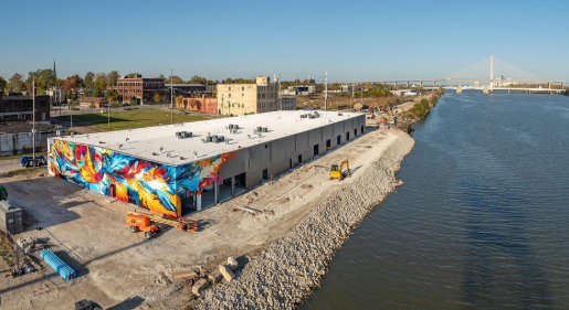 A large building by a river, with a colorful mural on one side. Construction equipment is nearby, and a bridge spans the river in the background.