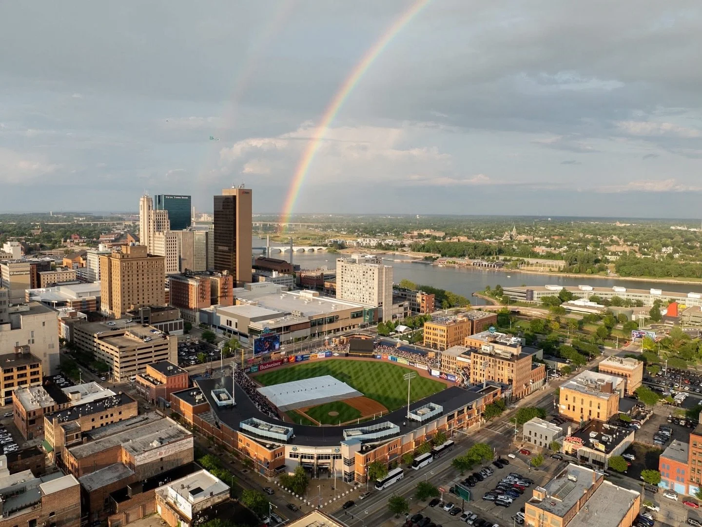 A cityscape with a baseball stadium in the foreground, tall buildings in the middle, a river with a bridge, and a rainbow in the sky above the city.