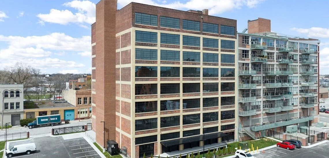 A multi-story office building with glass windows and brick exterior, next to a residential building with clear balconies, with parking lots and vehicles in the foreground, under a partly cloudy sky.