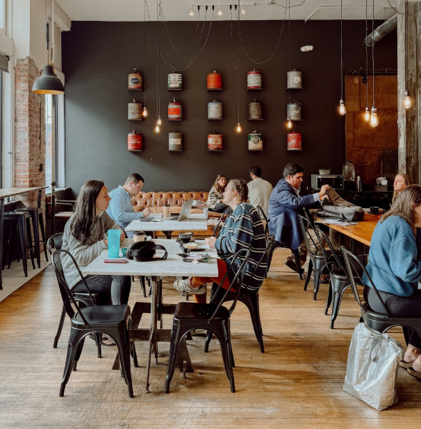 People working and dining in a modern, cozy cafe with black walls, hanging light bulbs, and decorative jars on the wall.