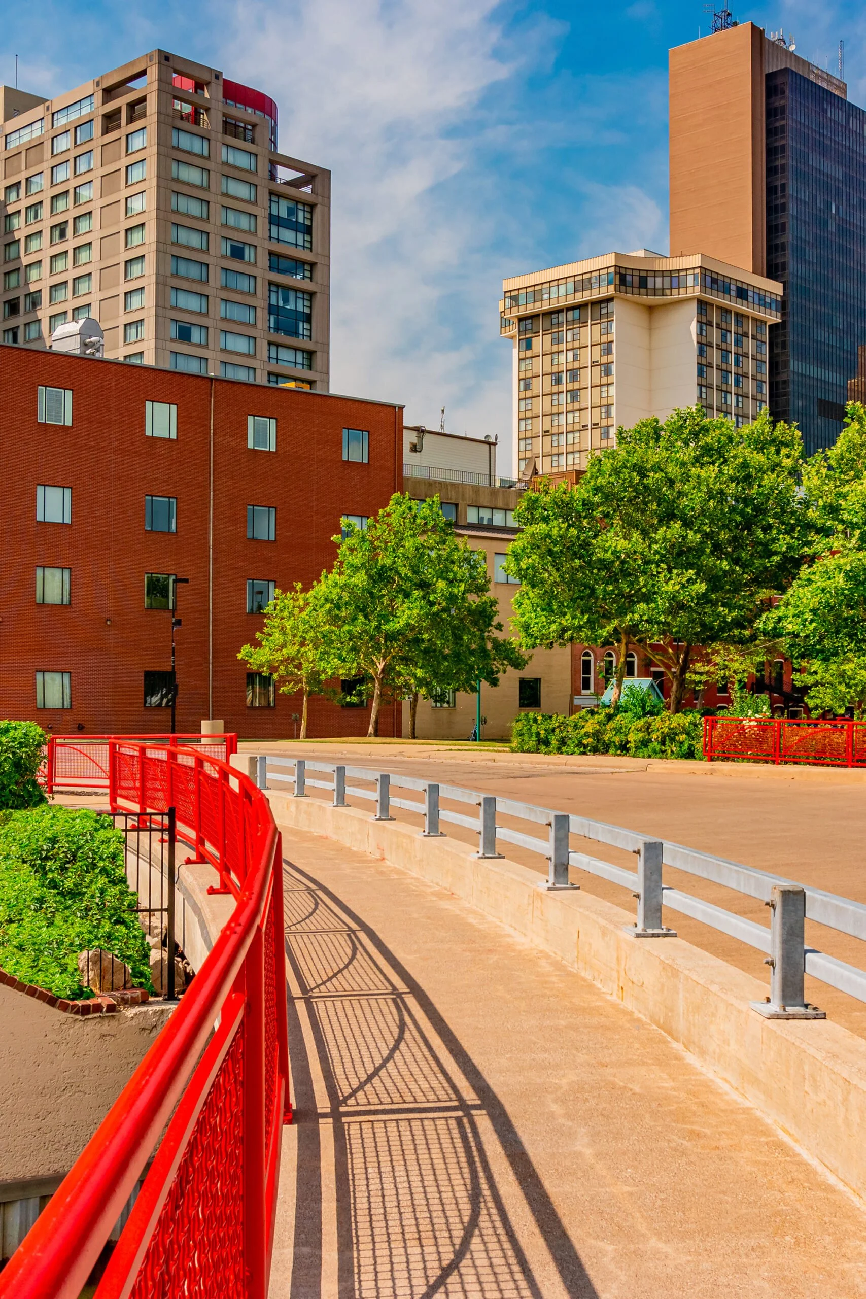 Cityscape with high-rise buildings, trees, and a pathway with red and gray railings in an urban park area.