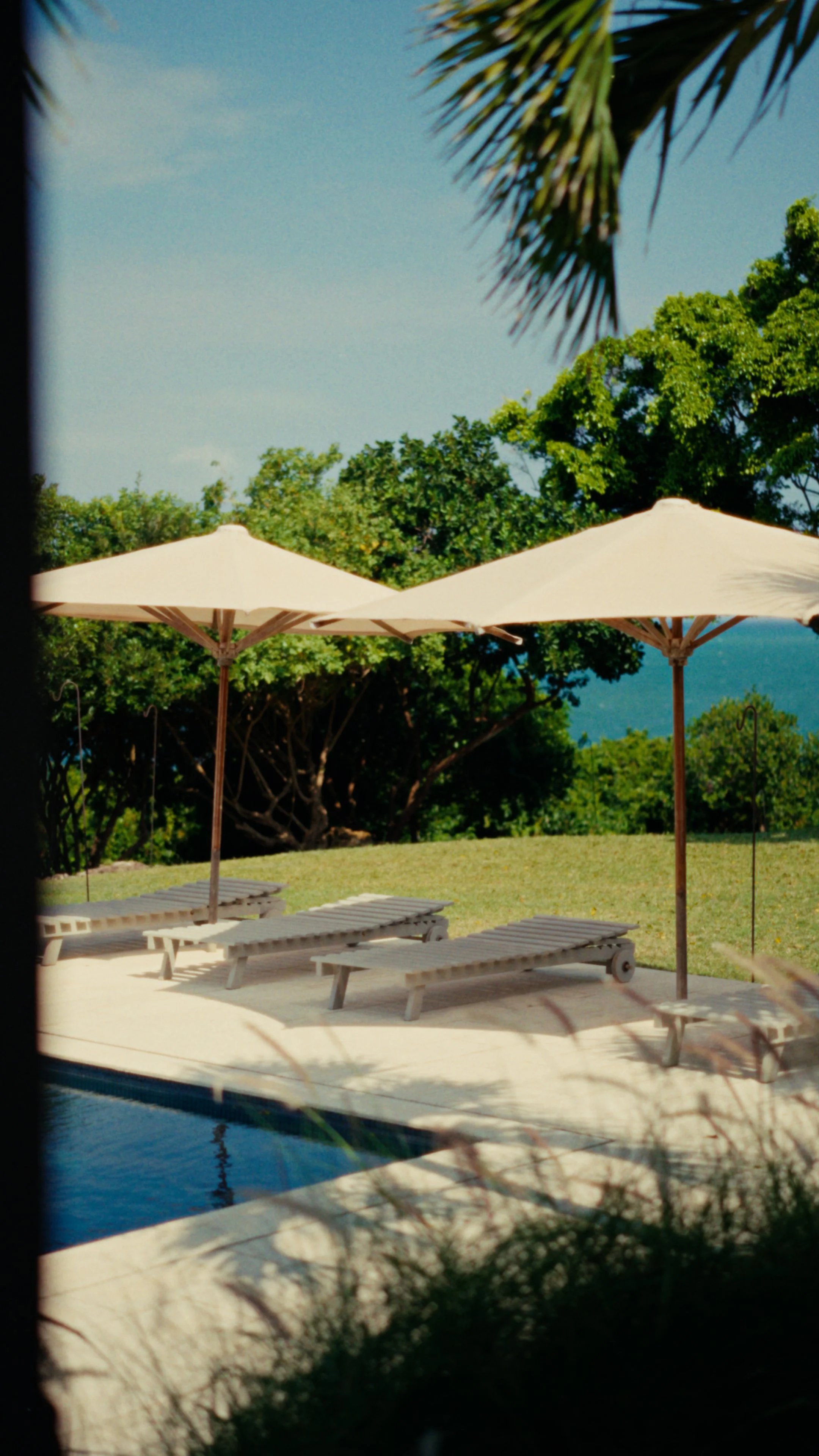 Poolside view with lounge chairs and large beige umbrellas, surrounded by green trees and clear blue sky.