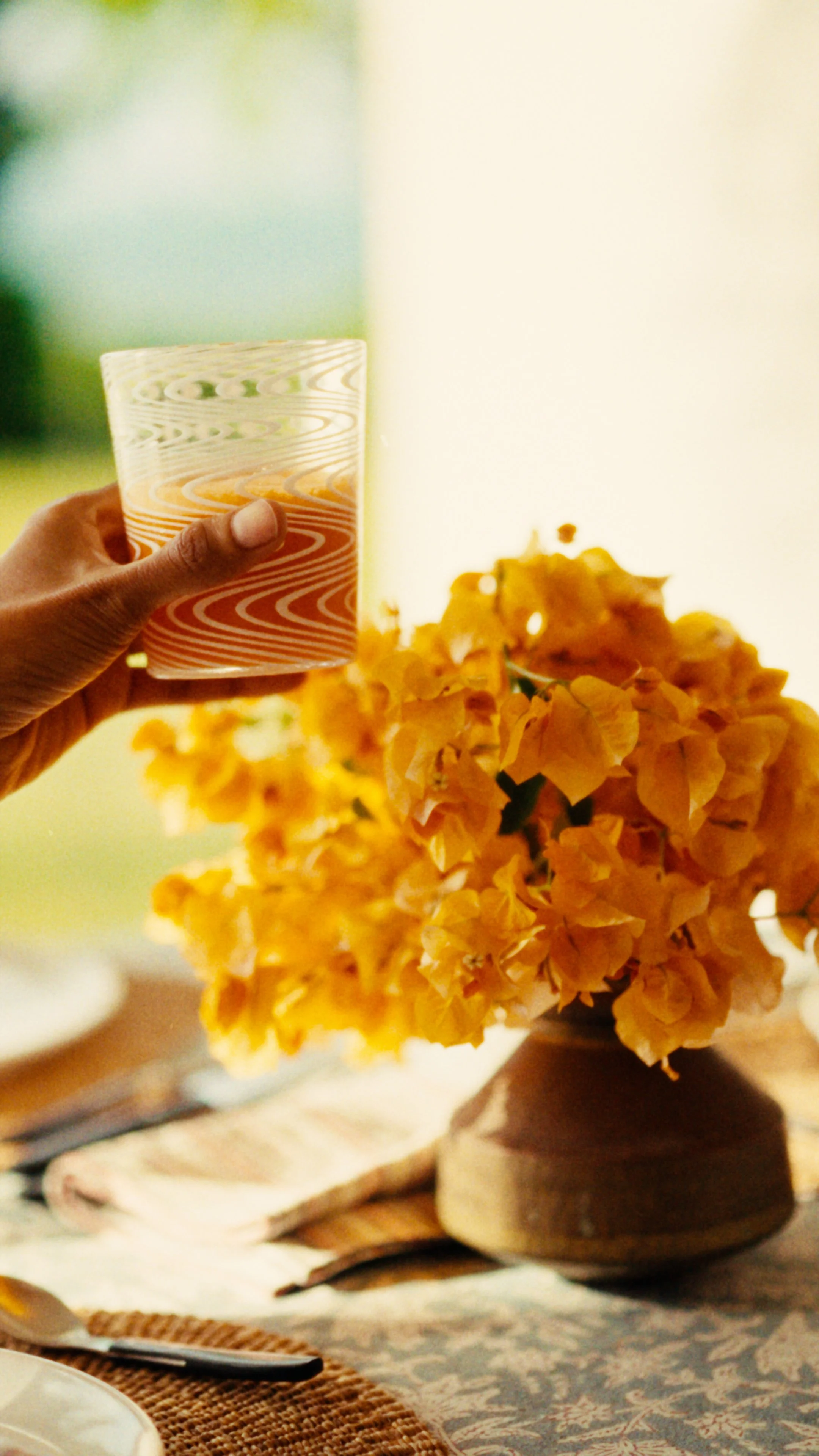 A person holding a glass of orange juice near a bouquet of orange flowers on a table setting.