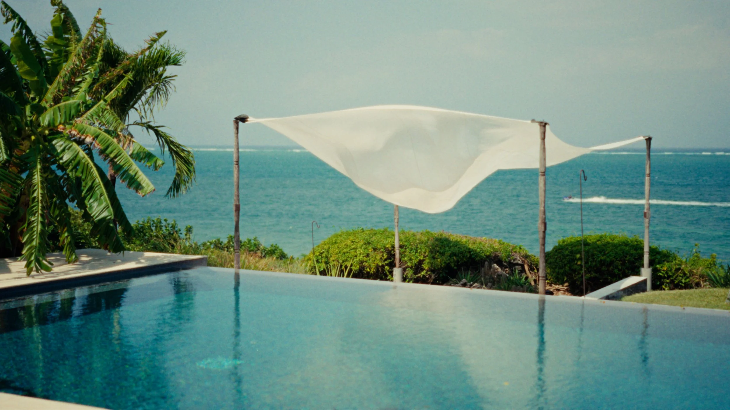 Beachside scene with a pool, green trees, and the ocean in the background, with white shade sails and boats on the water.
