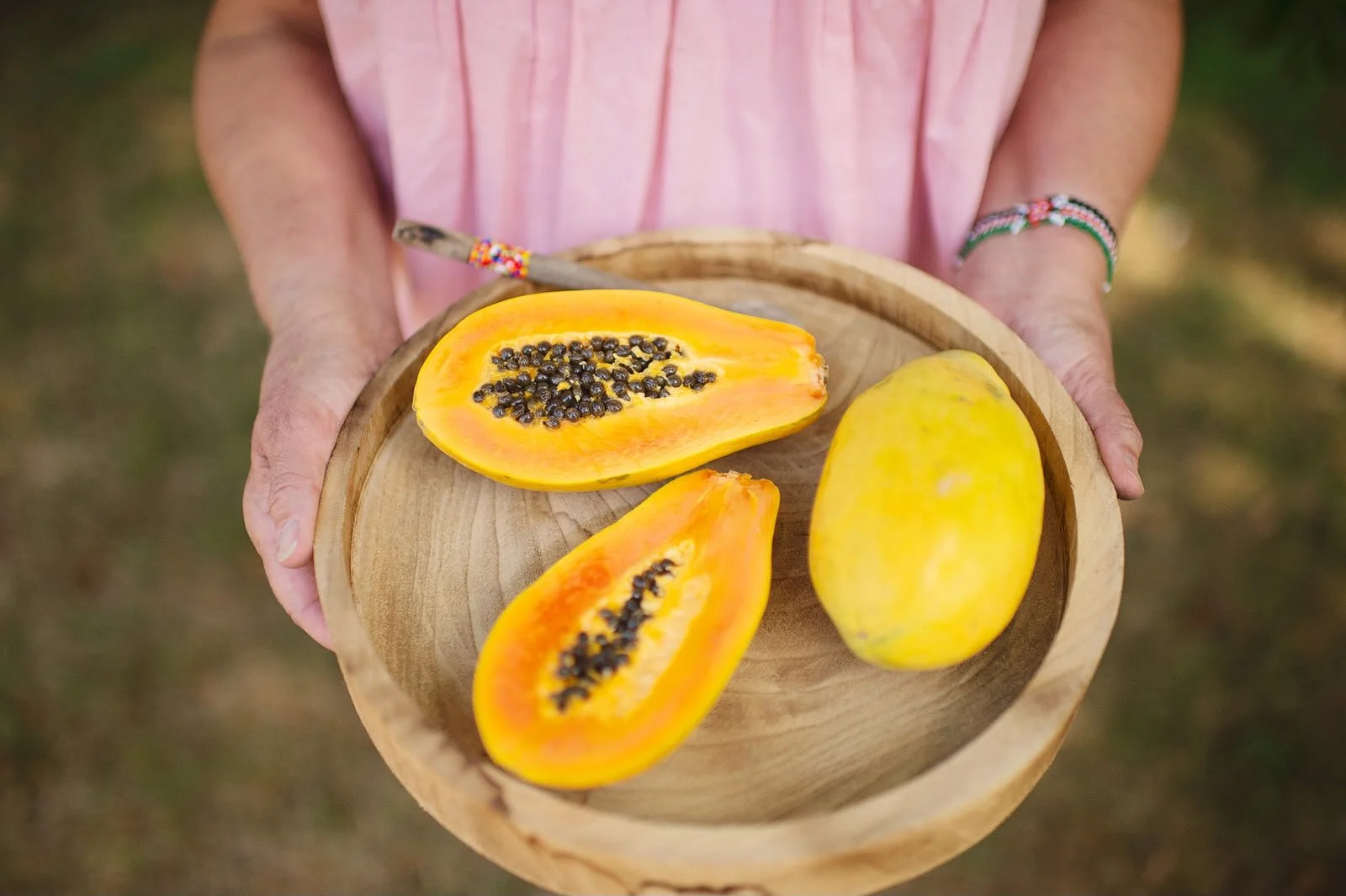 Person holding a round wooden tray with a halved yellow papaya showing black seeds and a whole yellow papaya.