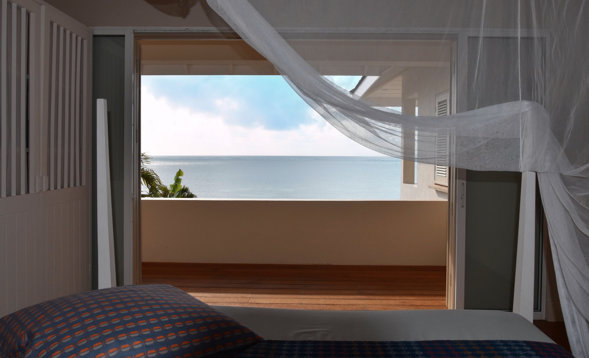 A bedroom with a view of the ocean through an open door leading to a balcony. The room has a bed with a patterned blanket and a pillow. Sheer curtains are flowing in the breeze.