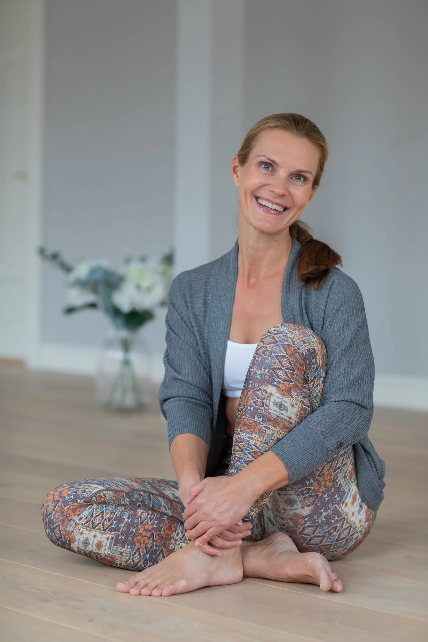 A woman with brown hair tied back, smiling, sitting cross-legged on the floor with her hands resting on her knees, indoors with a blurred floral vase in the background.