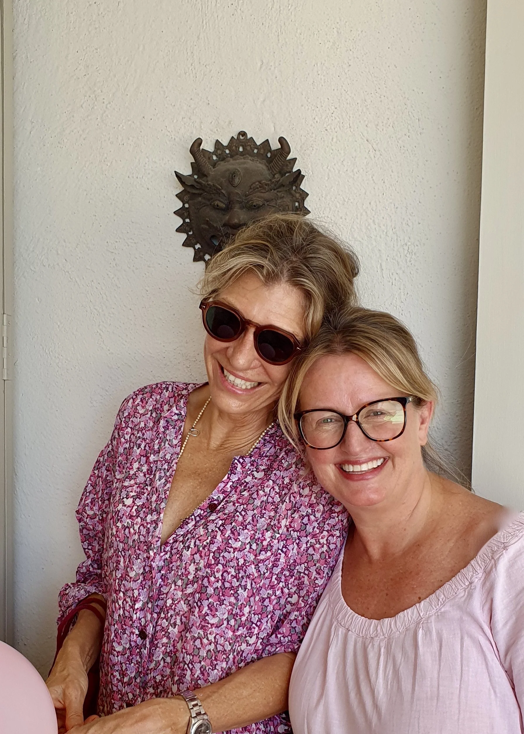 Two smiling women standing close together in front of a white wall with a decorative metal mask or sculpture of a creature with horns and multiple faces hanging on the wall behind them.