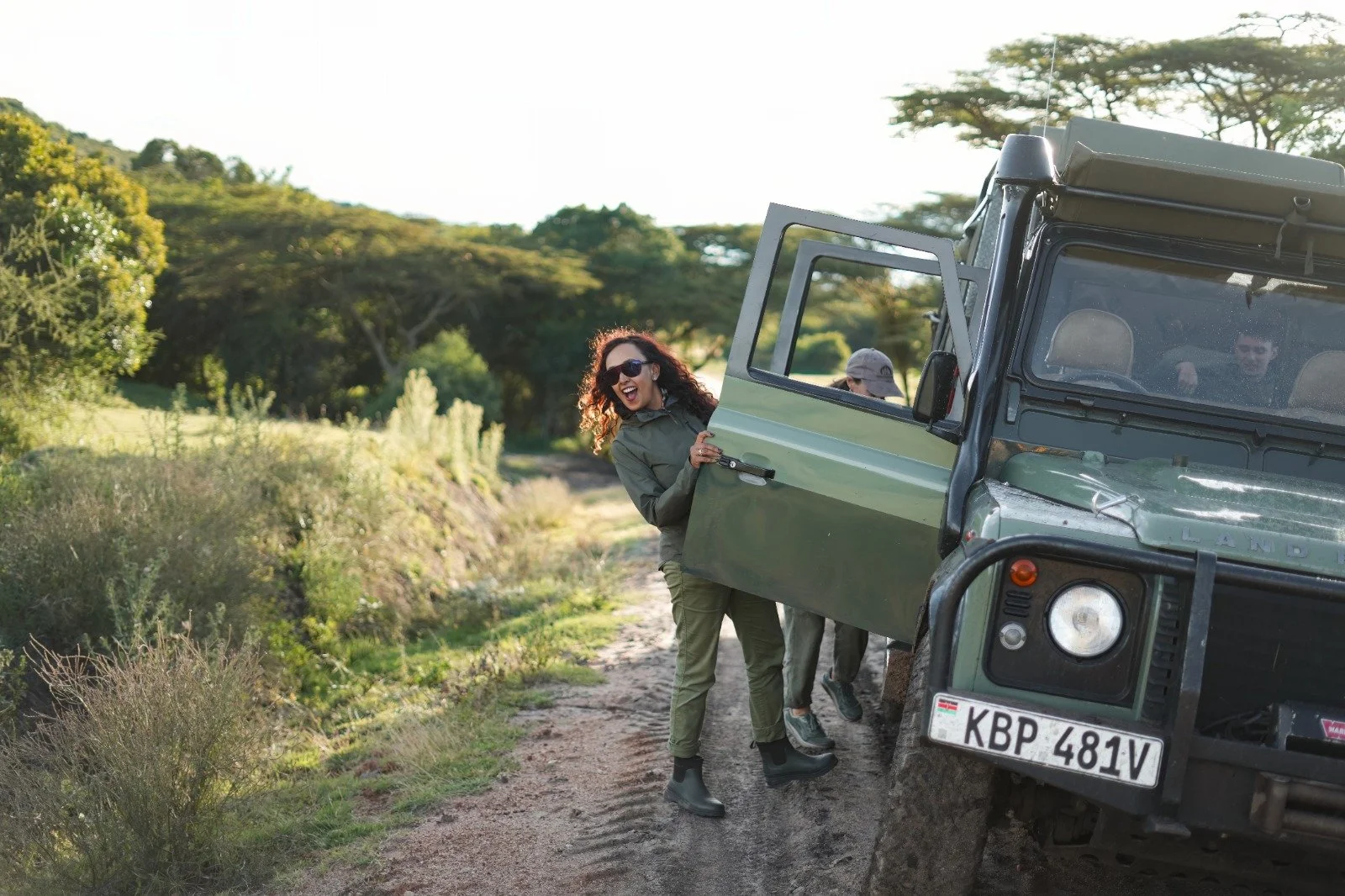 Woman in sunglasses and safari outfit smiling and leaning out of a safari vehicle on a dirt path surrounded by green trees and bushes.