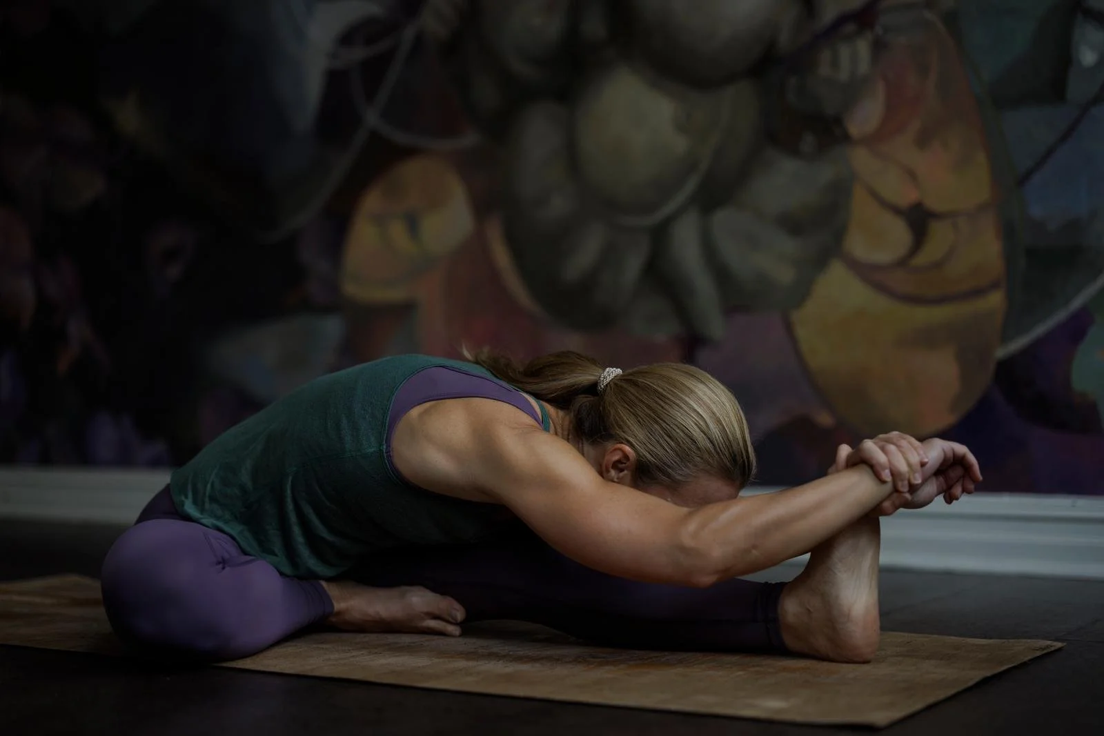 A woman practicing yoga in a seated forward bend pose on a mat indoors, with a large colorful mural of a cat in the background.