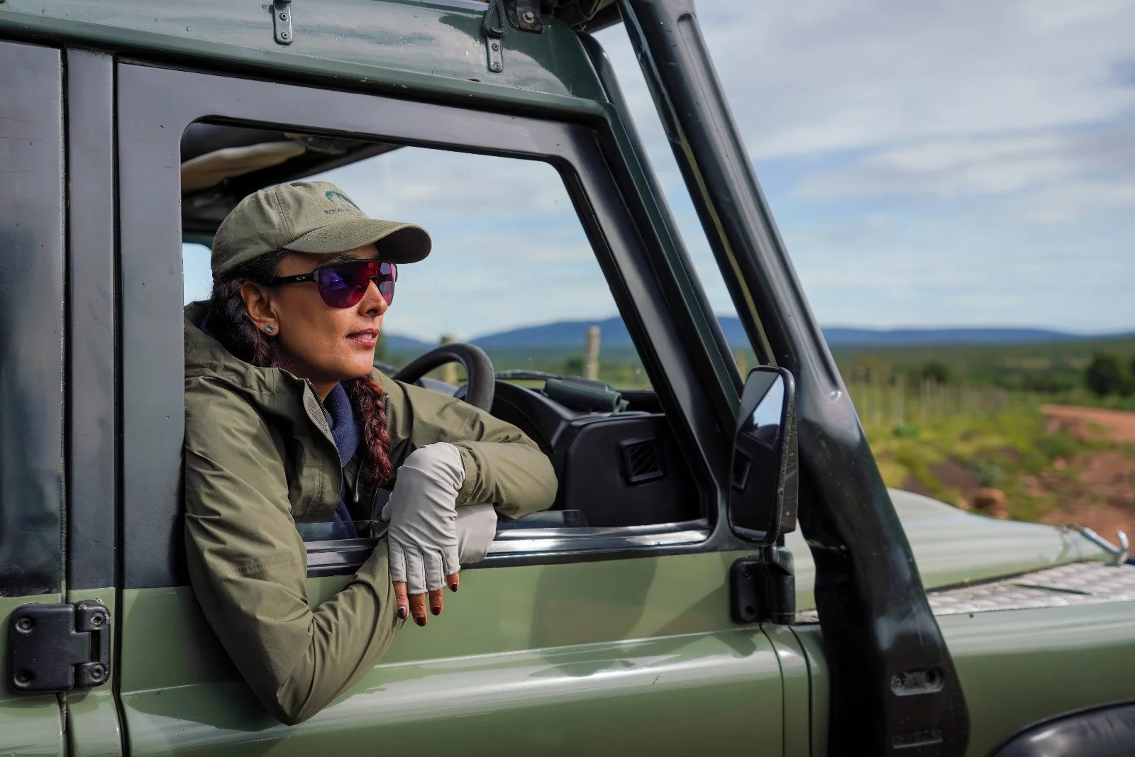 Woman in outdoor tactical gear and sunglasses sitting in a vehicle and looking into the distance with a landscape of fields and hills behind her.