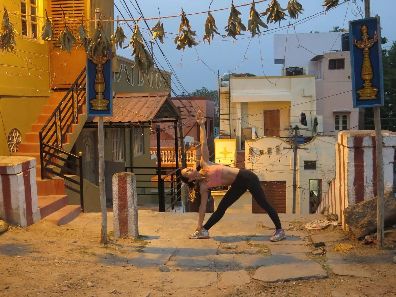 A woman in athletic clothing performing a yoga pose outdoors at sunset on a staircase in a neighborhood with colorful buildings and hanging leaf decorations.