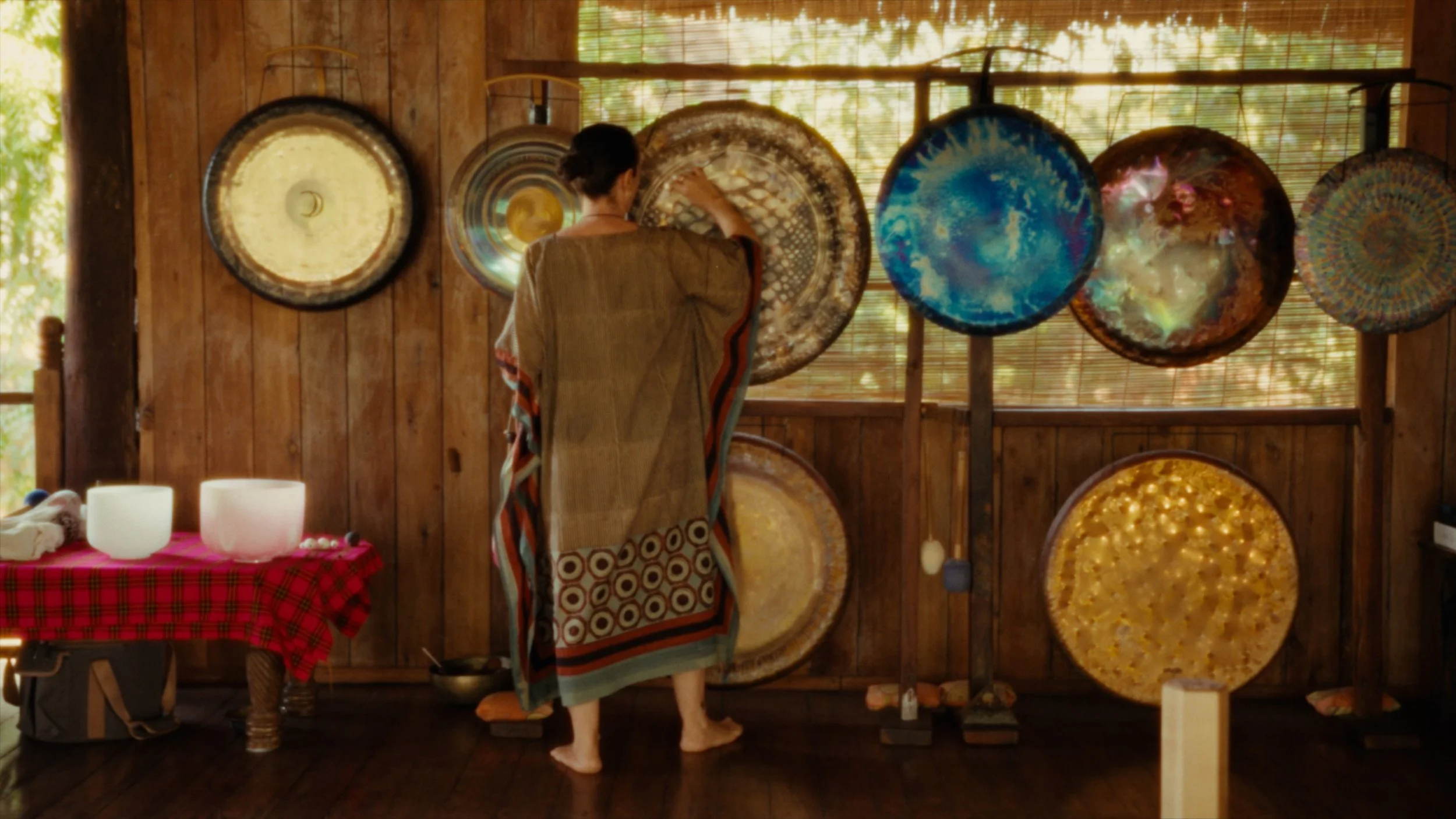 A woman dressed in a beige robe browsing large decorative drums hanging on a wooden wall in a rustic shop or studio. The drums are colorful and textured. There is a table with a red checkered cloth and white candles in the foreground.