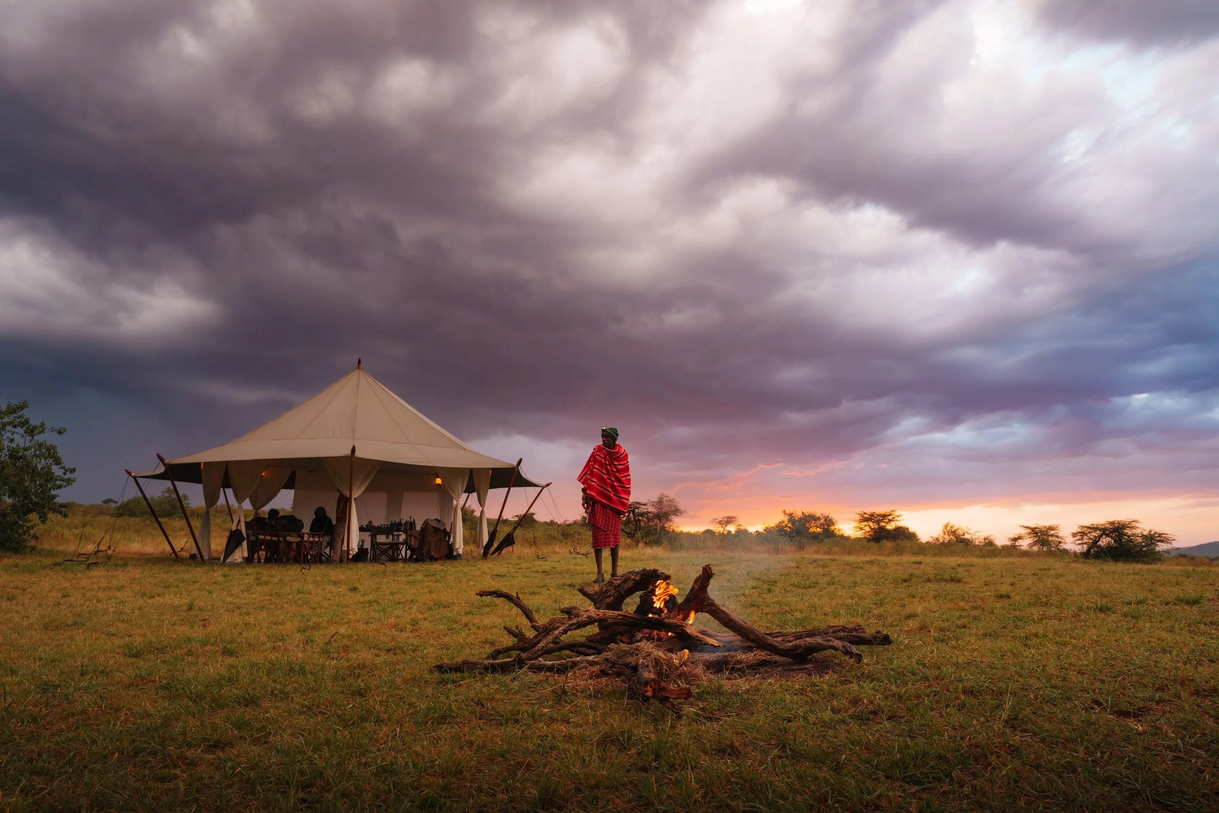 A person dressed in traditional Maasai clothing standing near a campfire in an open grassy landscape at sunset. There is a large beige tent with tables and people inside in the background under a dark, cloudy sky.