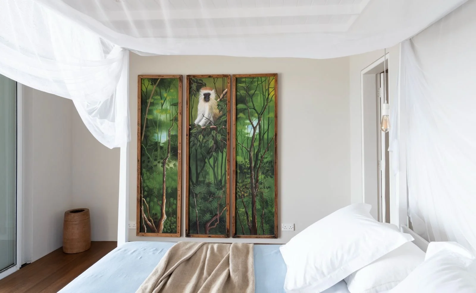 A bedroom with white bed linens, a three-panel rainforest-themed painting with a white lemur, a wicker basket in the corner, and white curtains draped around the room.