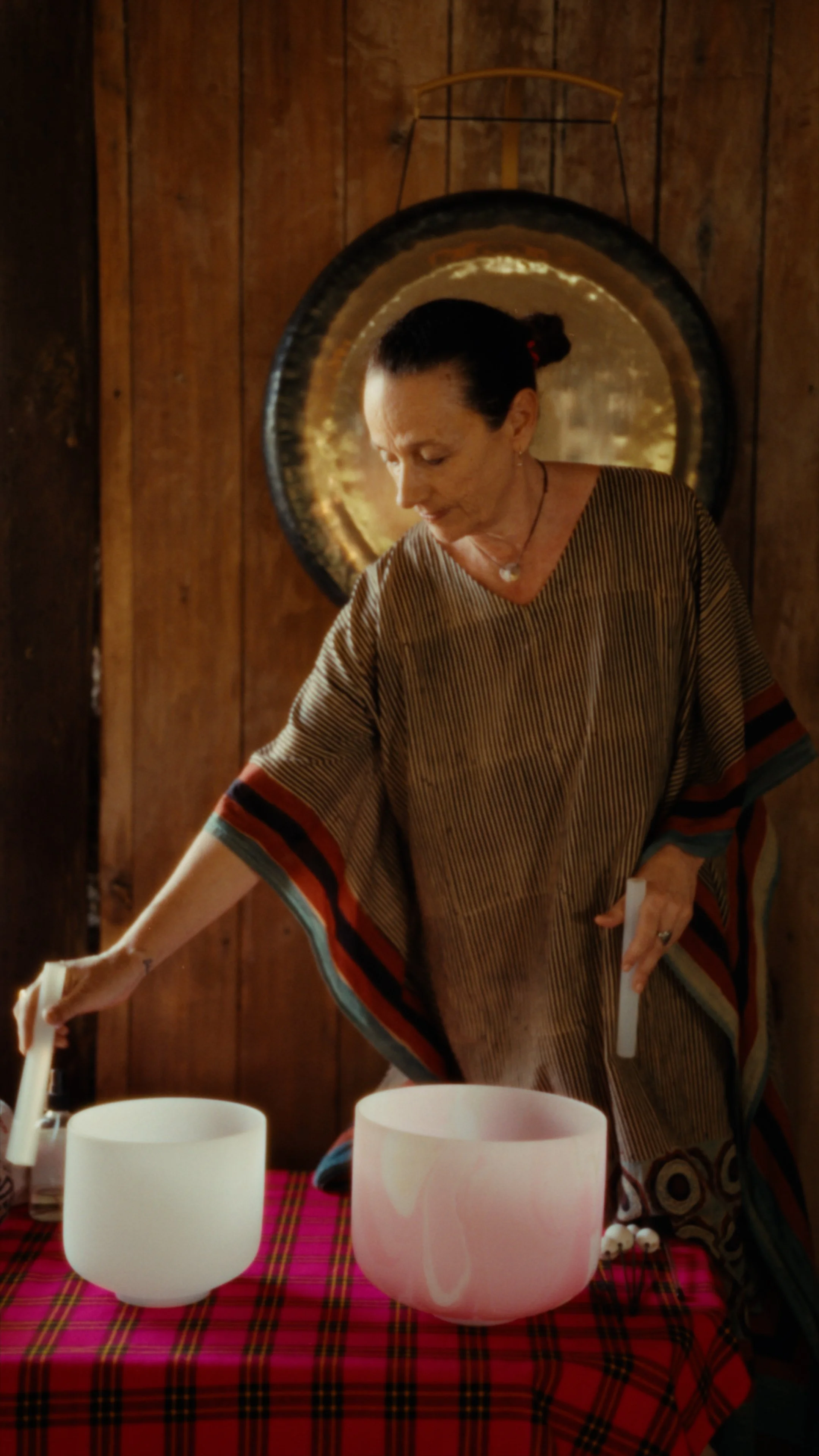 A woman with dark hair tied back, wearing a striped, multicolored poncho, plays singing bowls on a table covered with a red plaid cloth, against a wooden wall with a round decorative mirror behind her.