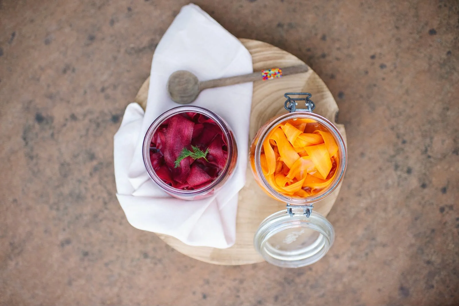 Two glass jars with pickled vegetables, one with red beets and the other with yellow peppers, on a round wooden tray with a white cloth and a small wooden pestle.