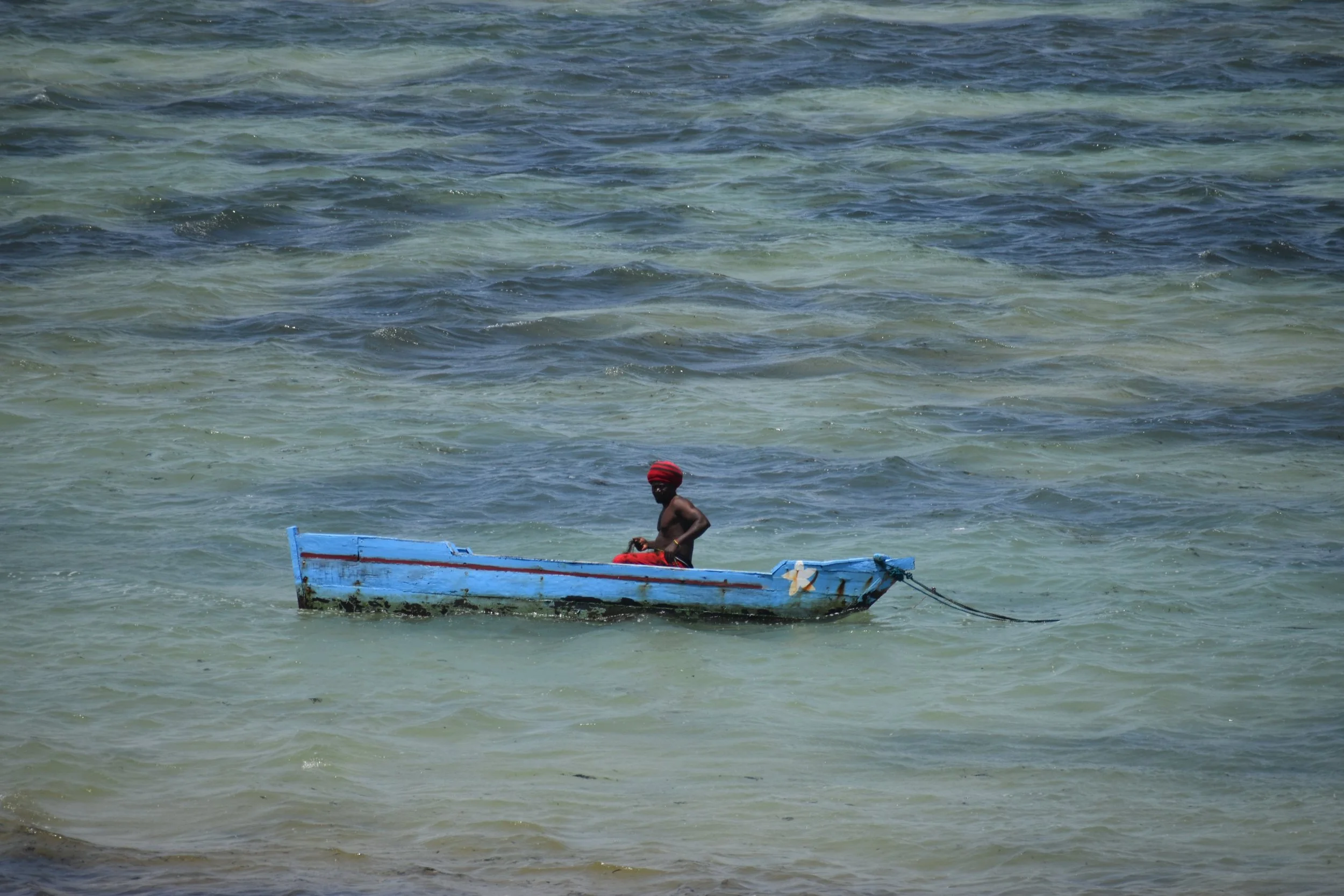 A person wearing a red cap on a small, weathered blue boat in the ocean near the shoreline.