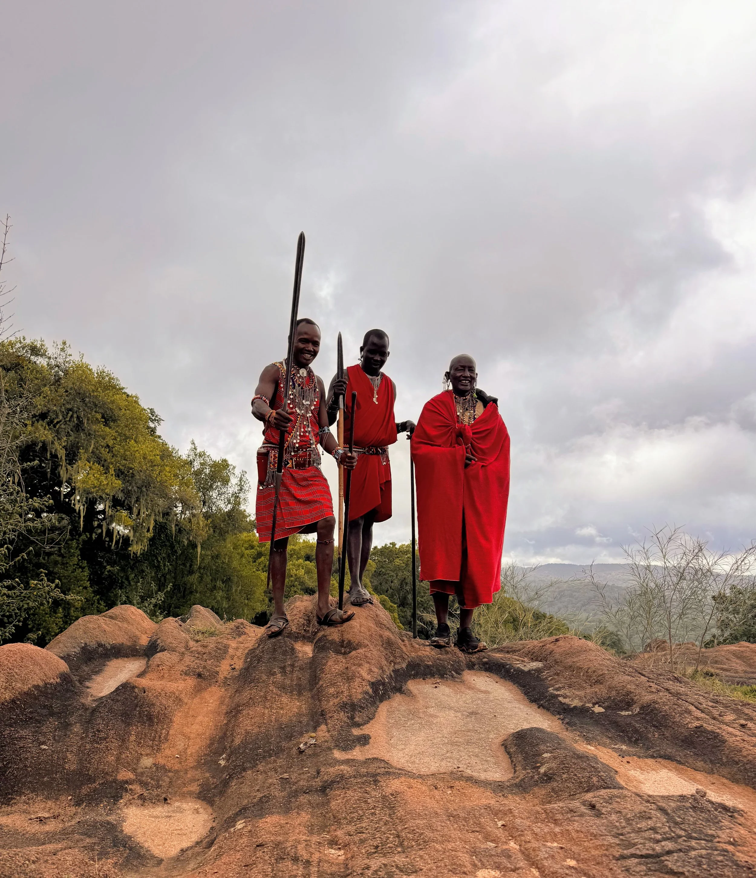 Three Maasai men wearing traditional red shukas and elaborate beaded jewelry, standing on a rocky hilltop against a cloudy sky, with lush trees in the background.