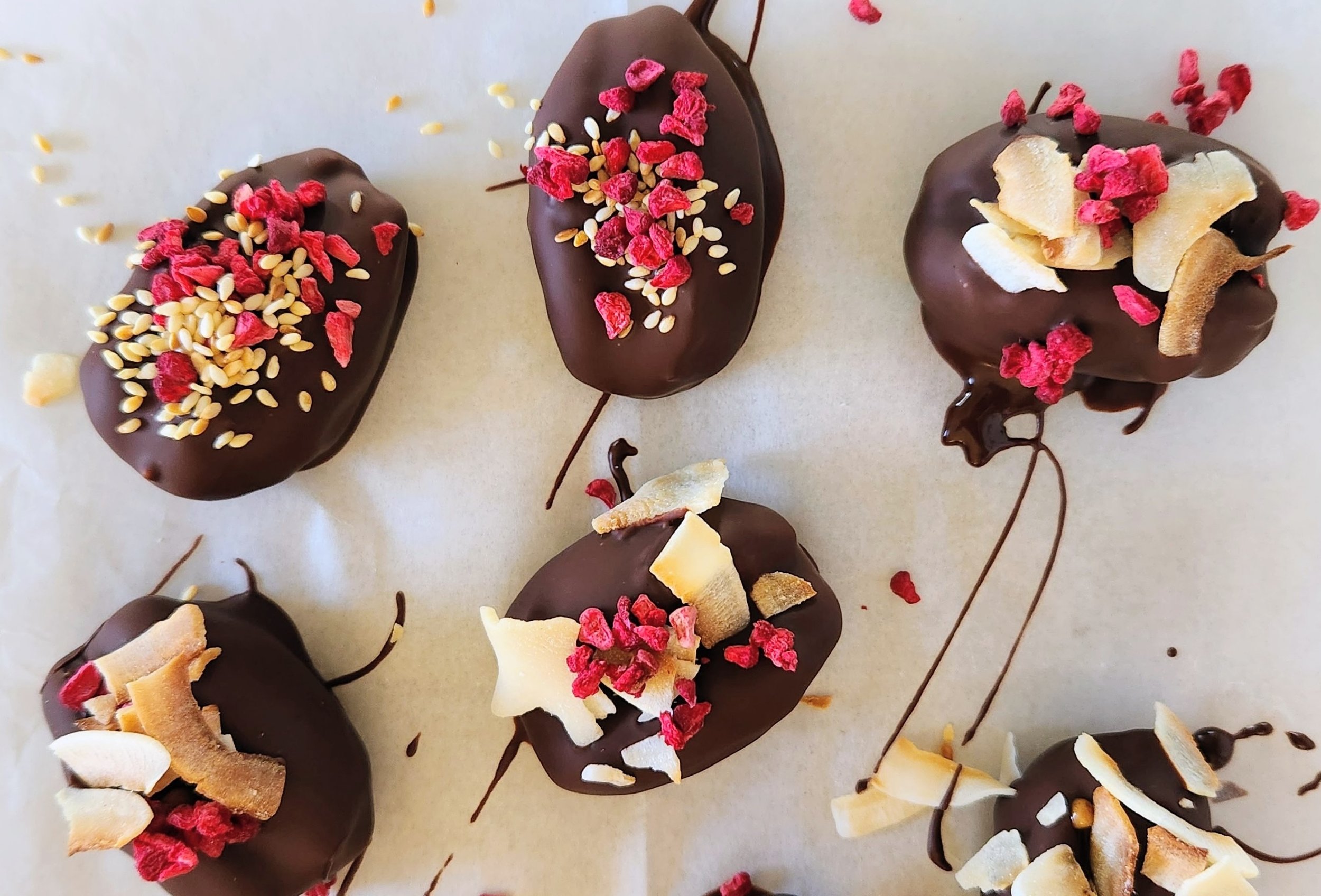 Chocolate-covered treats topped with dried berries, chopped nuts, and coconut flakes on white paper